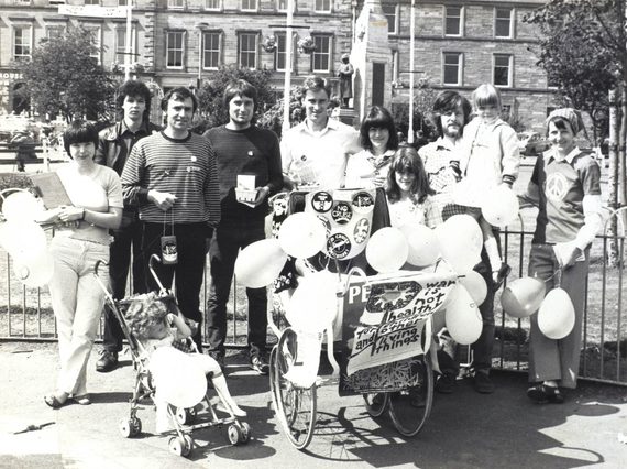 A black and white photo of a group of people standing in a city square holding balloons. There is a child's pram covered in protest signs and buttons.