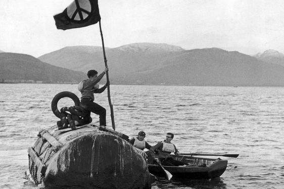 A black and white photo of a person holding a flag, standing on a submarine which is at the surface of a loch.