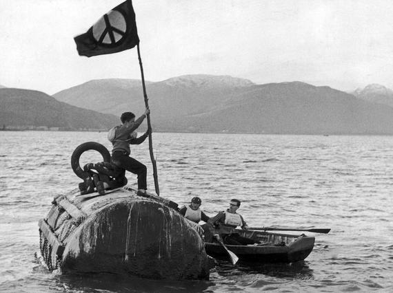 A black and white photo of a person holding a flag, standing on a submarine which is at the surface of a loch.