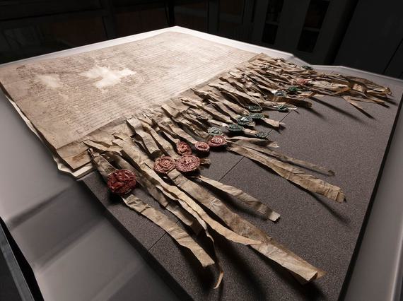 The Declaration of Arbroath laid out on a grey foam protective base set against a black background. The Declaration has dense Latin script across it with two small patches missing, and many strips dangling from the bottom affixed with green and red wax seals.