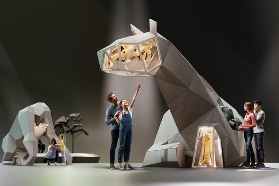 A man and young girl are standing in the centre of the image, pointing upwards at a giant, 3D sculpture of an animal. Other visitors are to the left and right, looking at the same sculpture, and the sculpture of a gorilla to the left