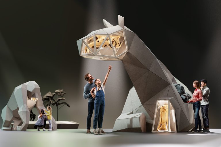 A man and young girl are standing in the centre of the image, pointing upwards at a giant, 3D sculpture of an animal. Other visitors are to the left and right, looking at the same sculpture, and the sculpture of a gorilla to the left
