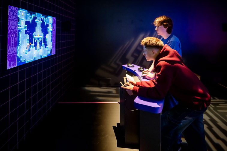 2 visitors to the Game On exhibition are holding consoles and playing bomberman.