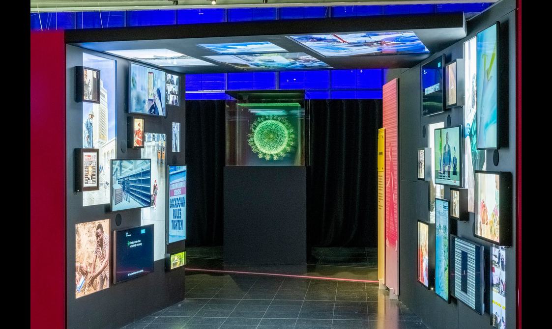 An exhibition display. At the centre of the image is an artwork on top of a black plinth. It looks like a light installation in the shape of a neon green virus cell.