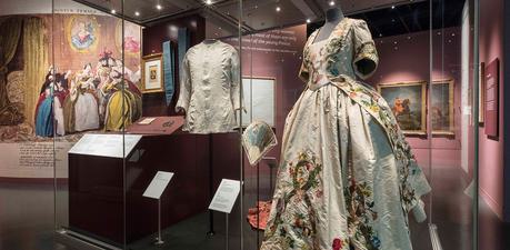 Garments on display in a wide museum case. On the left is a white long-sleeved shirt and on the right is a floral dress with wide skirt.