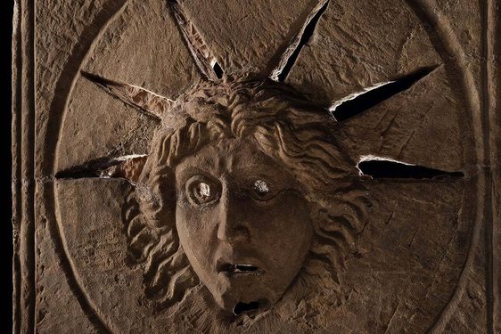 A Roman stone altar with the face of the Roman god Sol carved into it. Rays of light pass through slits around the head of Sol.