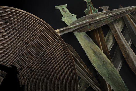 An array of bronze age weapons including a brown shield with a hole in the centre, a dark grey spearhead and a stack of green and grey short swords against a black background.