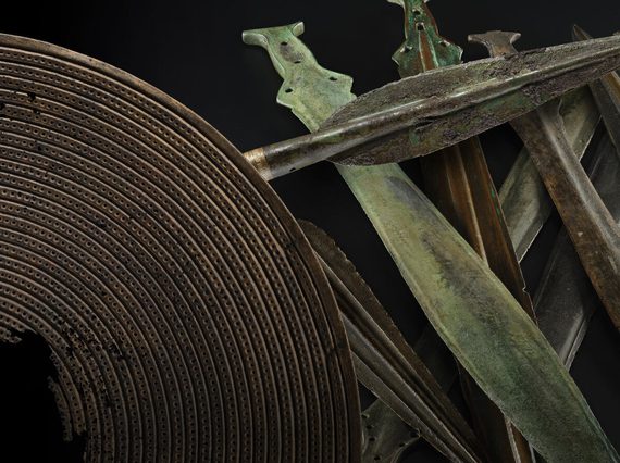 An array of bronze age weapons including a brown shield with a hole in the centre, a dark grey spearhead and a stack of green and grey short swords against a black background.