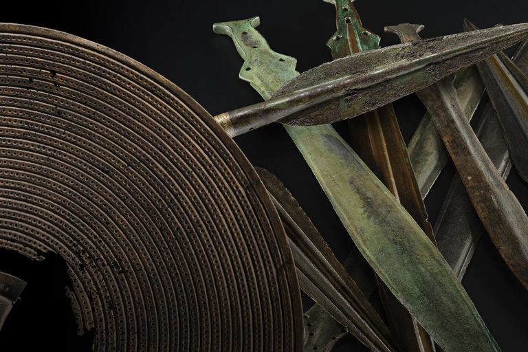 An array of bronze age weapons including a brown shield with a hole in the centre, a dark grey spearhead and a stack of green and grey short swords against a black background.