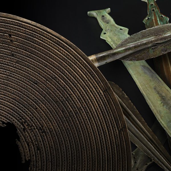 An array of bronze age weapons including a brown shield with a hole in the centre, a dark grey spearhead and a stack of green and grey short swords against a black background.