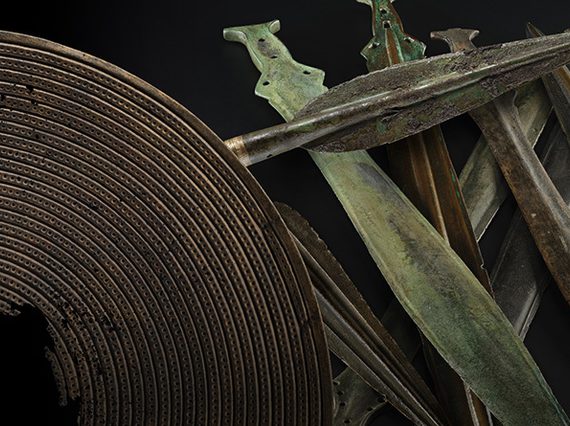 An array of bronze age weapons including a brown shield with a hole in the centre, a dark grey spearhead and a stack of green and grey short swords against a black background.