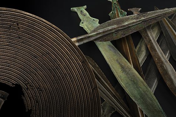 An array of bronze age weapons including a brown shield with a hole in the centre, a dark grey spearhead and a stack of green and grey short swords against a black background.