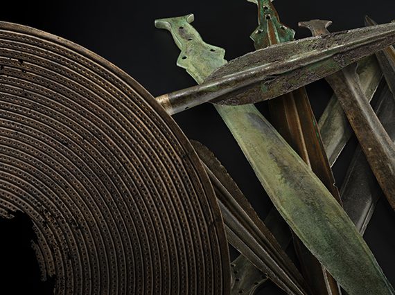 An array of bronze age weapons including a brown shield with a hole in the centre, a dark grey spearhead and a stack of green and grey short swords against a black background.