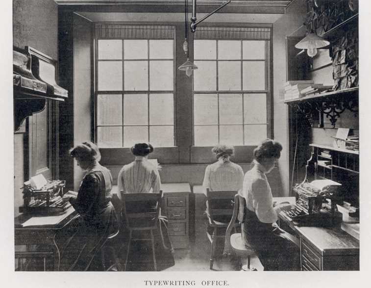 A black and white photograph of four typists working at office desks.