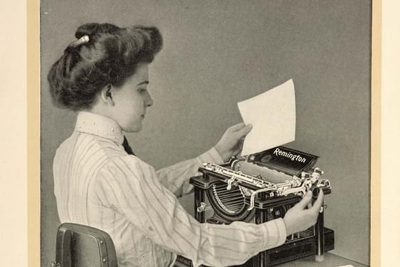 A black and white photograph of a seated typist inserting paper into a typewriter.