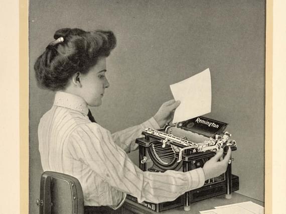 A black and white photograph of a seated typist inserting paper into a typewriter.