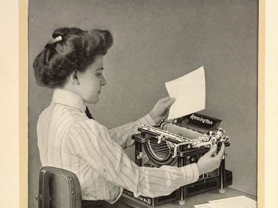 A black and white photograph of a seated typist inserting paper into a typewriter.