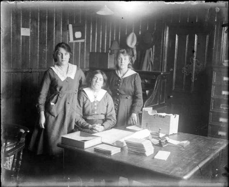 A black and white photograph of three clerical workers behind a desk in an early 20th century office.
