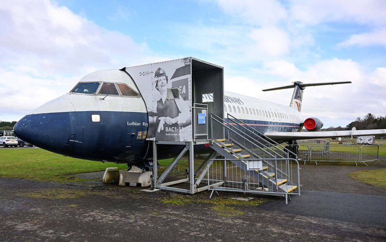 A white and blue commercial aricraft on display on an airfield. A set of stairs lead up to the entrance of the aircraft.