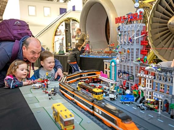 A parent and two young children standing at a table displaying a model Lego city and train models.