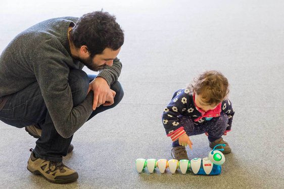 An adult and child crouch over a colourful caterpillar robot toy with big green lights for eyes.
