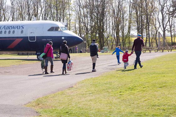 Adults and children walking along a tarmac path in an airfield. There is a parked British Airways aircraft in the distance next to some picnic tables.