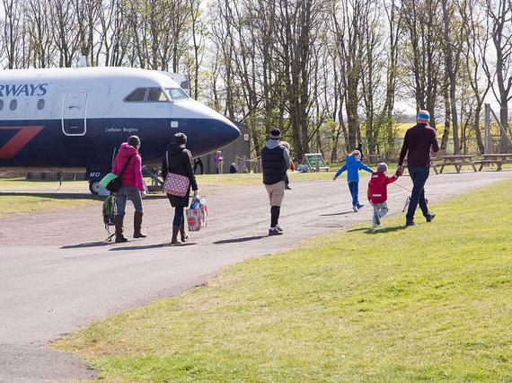 Adults and children walking along a tarmac path in an airfield. There is a parked British Airways aircraft in the distance next to some picnic tables.