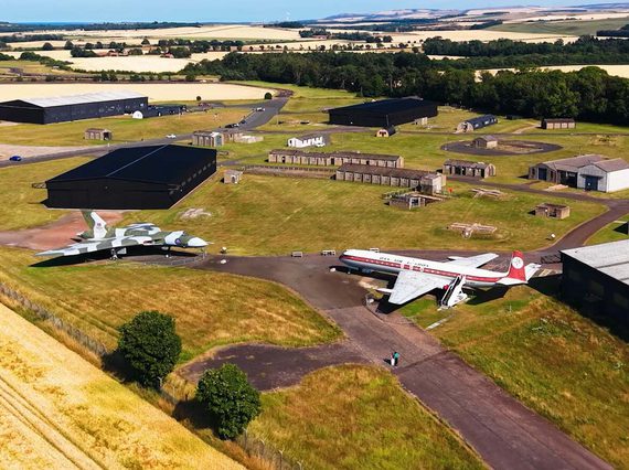 A historic war airfield with different buildings and hangars. Several aircraft are on display in the airfield.