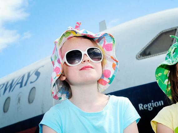 Two children in sunglasses and hats look up with a British Airways plane behind them.