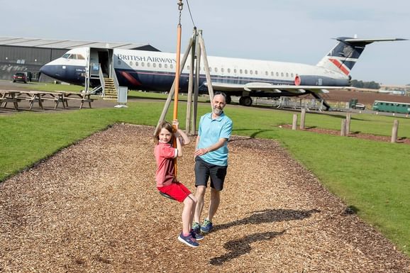 An adult pushing a child along a zipline on an assault course. There is a British Airways aircraft parked in the background.