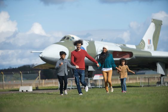 A family of four holding hands, running in a grassy field, with a large Vulcan aircraft in the background.