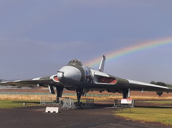 A Vulcan aircraft stationed in an airfield outside a black aviation hangar. There is a rainbow in the sky in the background.