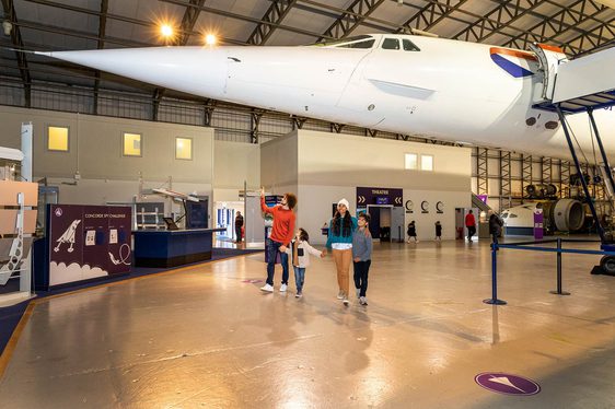 A family of four in a museum walking under the nose of a Concorde aeroplane.
