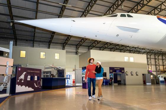 A couple in a museum walkiing under the nose of a Concorde aeroplane.