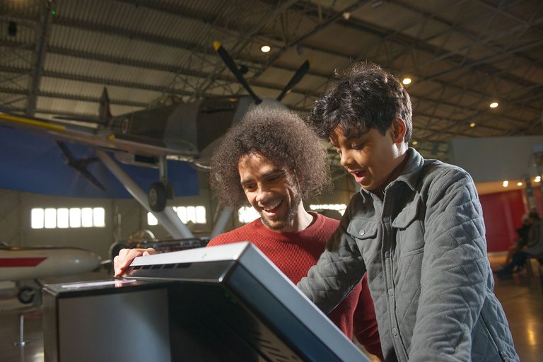 A parent and child in an aircraft hangar interacting with a screen, with a large aircraft in the background.