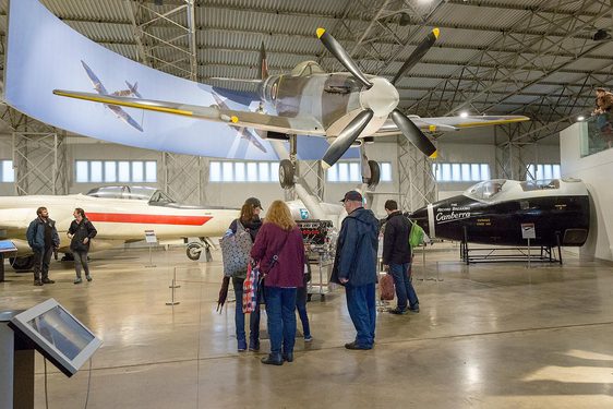 A group of visitors look up at a Spitfire aircraft in an aviation hangar.