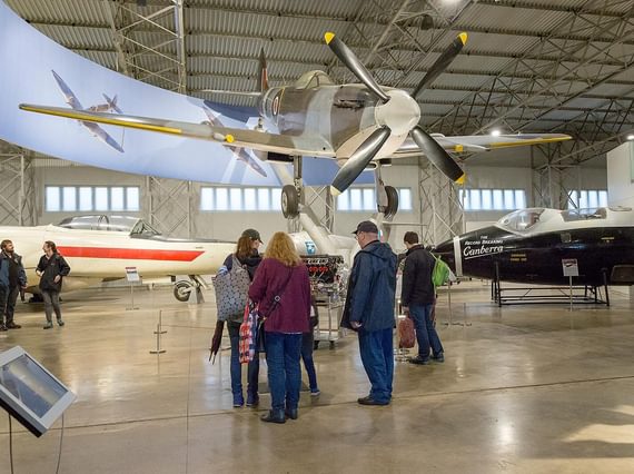 A group of visitors look up at a Spitfire aircraft in an aviation hangar.