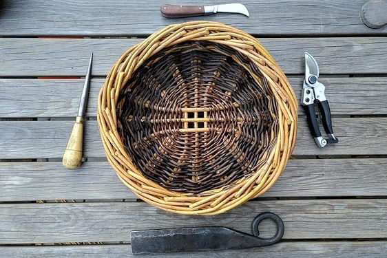 A round willow basket pictured from above surrounded by four tools for willow making