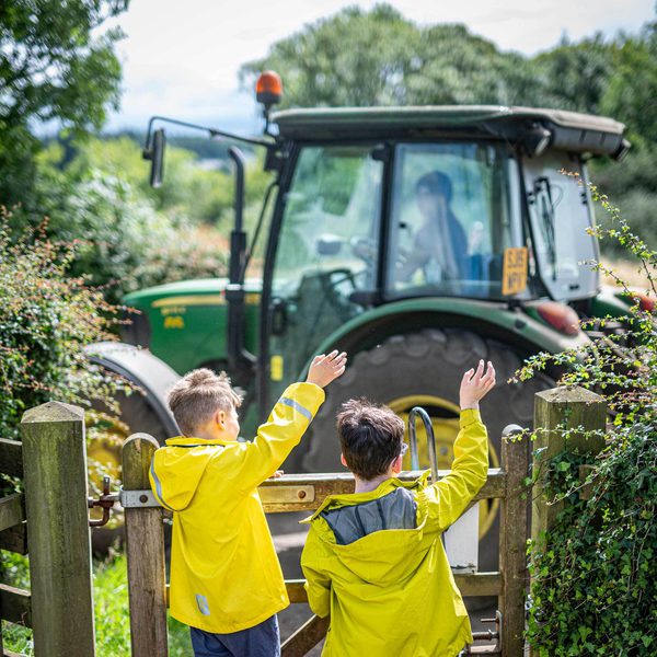 Two children in yellow jackets waving at a green tractor.