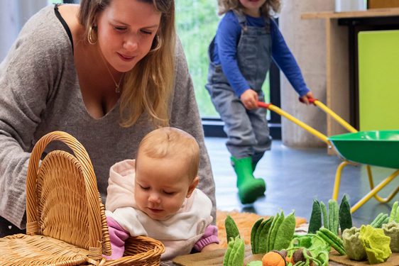 A parent and child play with soft vegetable toys in a playroom. Another child is in the background pushing a wheelbarrow.
