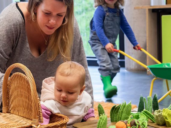 A parent and child play with soft vegetable toys in a playroom. Another child is in the background pushing a wheelbarrow.