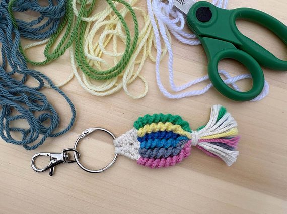A colourful woven fish keyring on a table next to strands of thread and a pair of scissors.