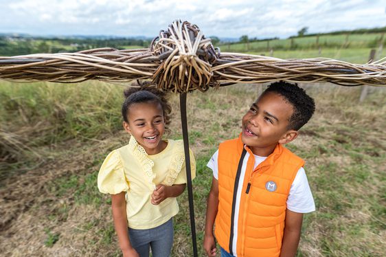 Two children standing underneath a bird sculpture made of willow.