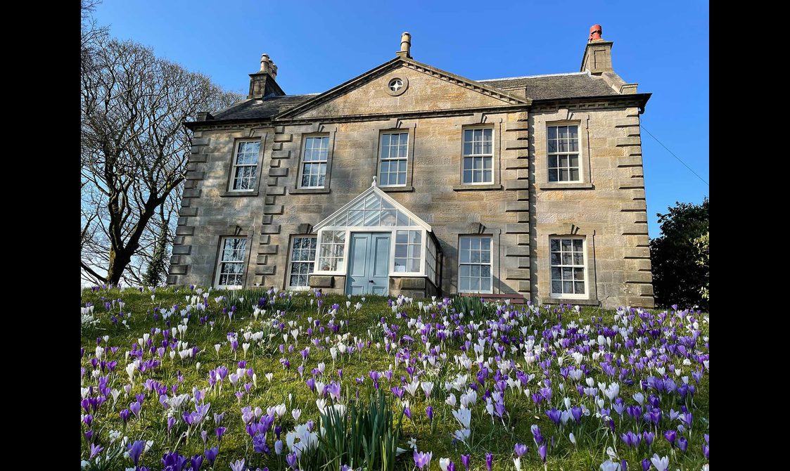 A large brick farmhouse with white and purple flowers in the garden.
