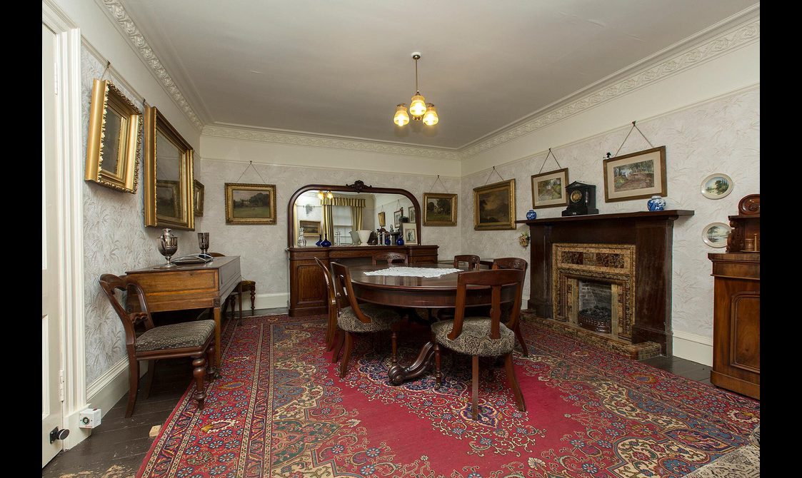 The interior of a vintage dining room, with ornate wooden furniture and framed portraits on the walls.