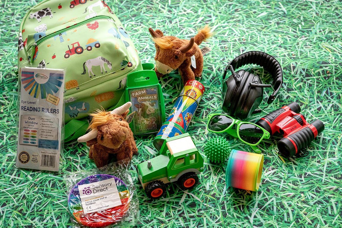 A green backpack with sensory items next to it, including ear defenders, binoculars, sunglassesm and fluffy highland cow toys.
