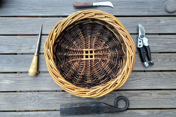 A brown willow basket surrounded by basket making tools on wooden decking.