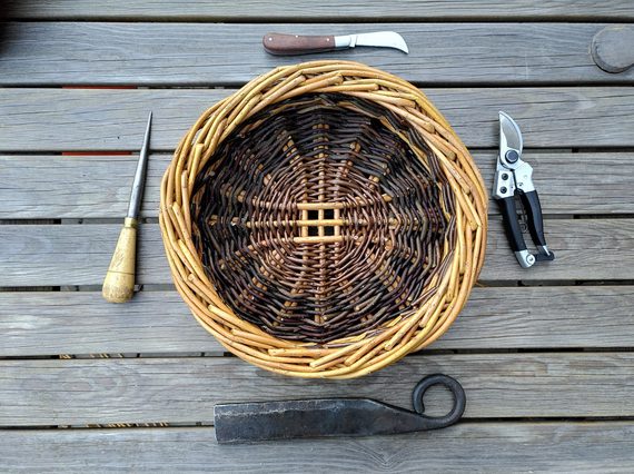 A brown willow basket surrounded by basket making tools on wooden decking.