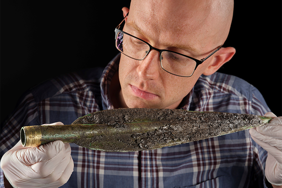 Man with glasses and white gloves hold a Bronze Age spearhead close to his face.
