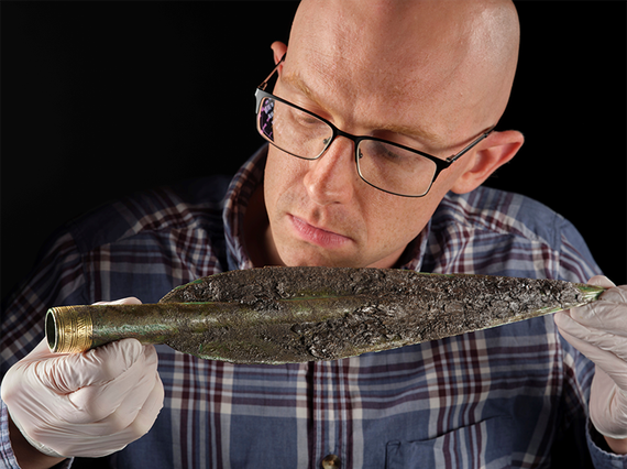 Man with glasses and white gloves hold a Bronze Age spearhead close to his face.
